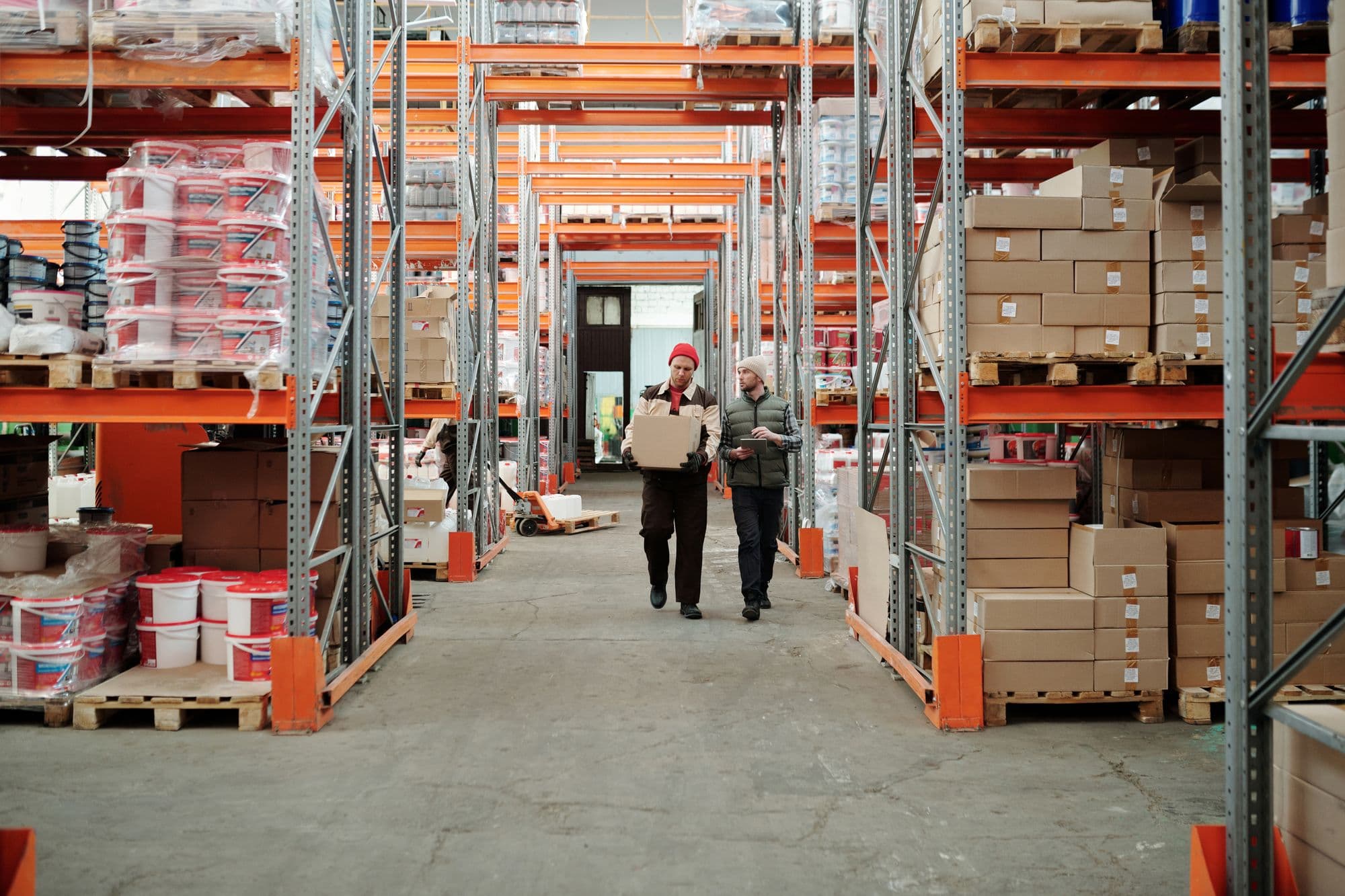 Two warehouse workers walking down an aisle between tall pallet racks, one carrying a box and one holding a tablet.